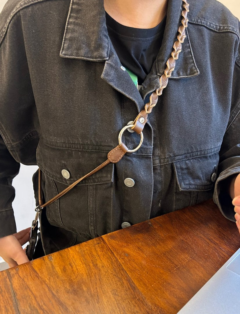 Person wearing black denim jacket with braided brown leather phone sling across chest at wooden table