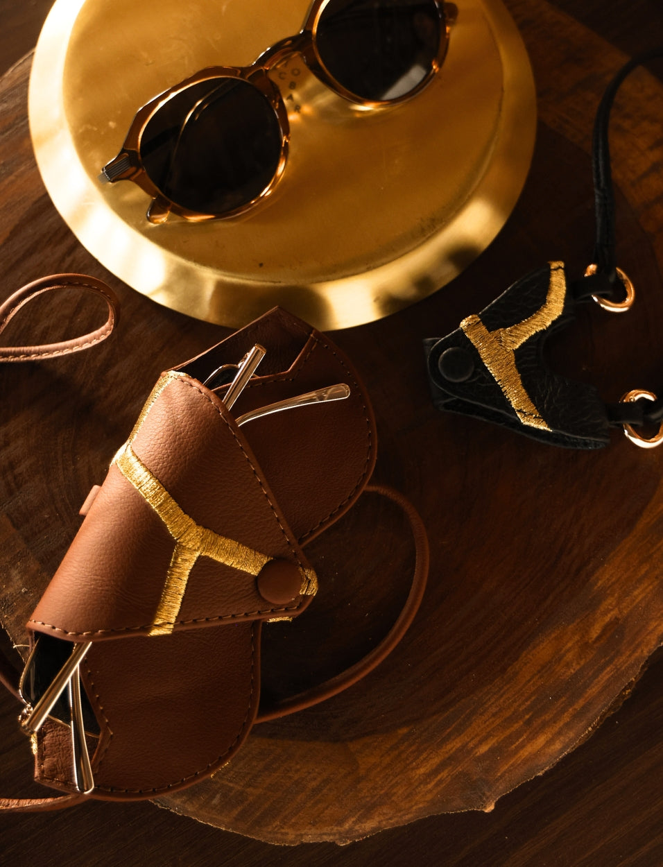 Brown and black leather eyeglass cases with gold embroidery, sunglasses on gold plate, wooden surface
