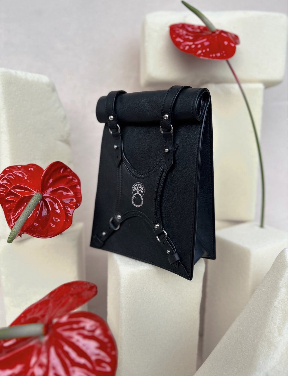 Black leather handbag with silver studs and strap details on white foam blocks with red anthurium flowers