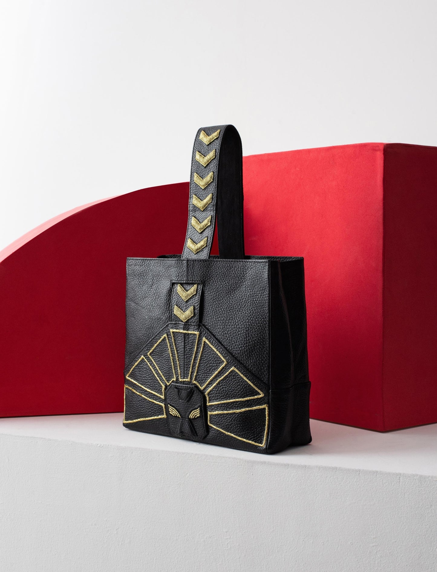 Black leather tote bag embossed with gold geometric patterns and arrows handle, against red and white background
