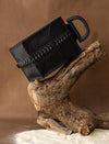 Black leather handbag with woven detail displayed on rustic wooden branch and beige fabric