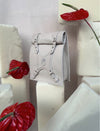 Beige leather handbag with buckle straps, displayed among cream foam blocks and red anthurium flowers
