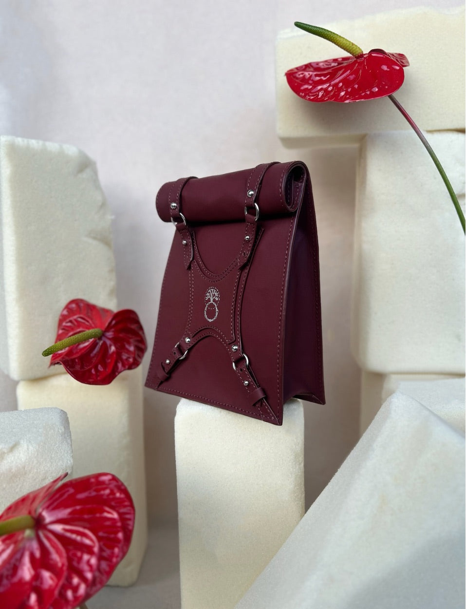 Maroon leather handbag with silver buckles displayed on white foam blocks with red anthurium flowers