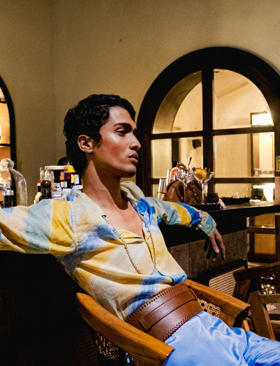 Man wearing a colorful tie-dye shirt and a wide tan leather waist belt, seated in a wooden chair indoors