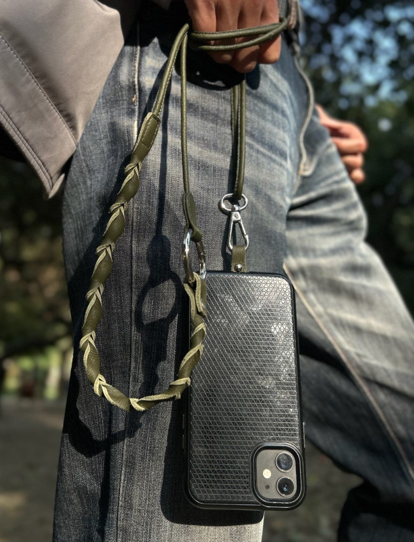 Hand holding a black textured phone case with an olive green leather upcycled lanyard against blue jeans outdoors