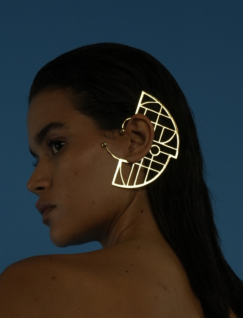 Profile of woman with sleek dark hair wearing large gold geometric metal ear cuff against blue background
