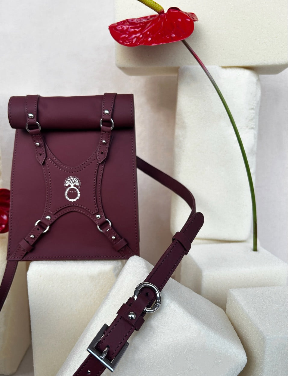Maroon leather backpack with silver hardware and a rolled top, displayed on white foam blocks with a red anthurium flower