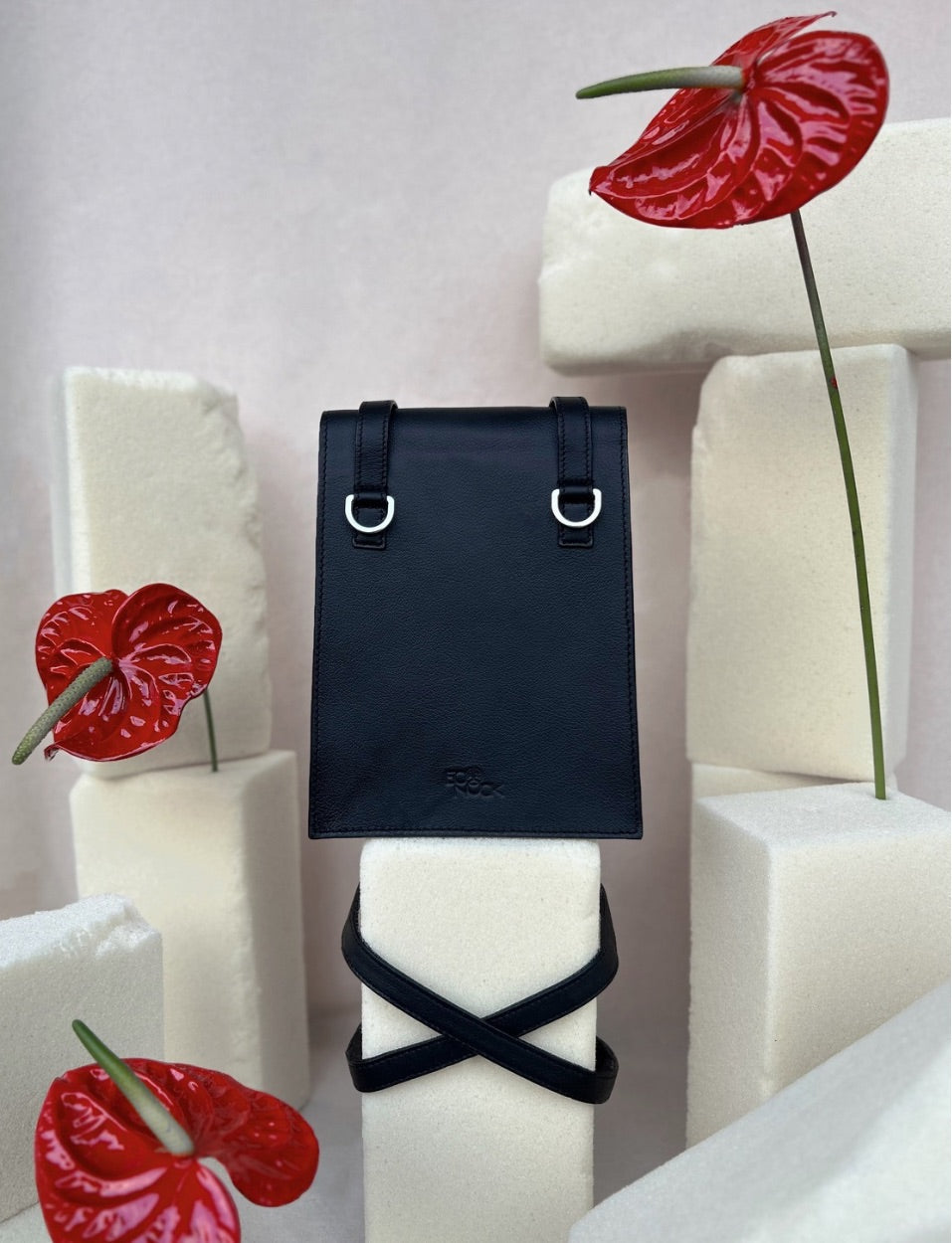 Black leather backpack displayed on white foam blocks with red anthurium flowers in minimalist studio