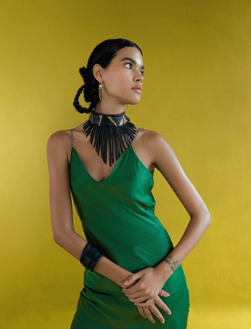 Woman in green dress wearing black fringe choker, gold earrings, and black bracelets against yellow background