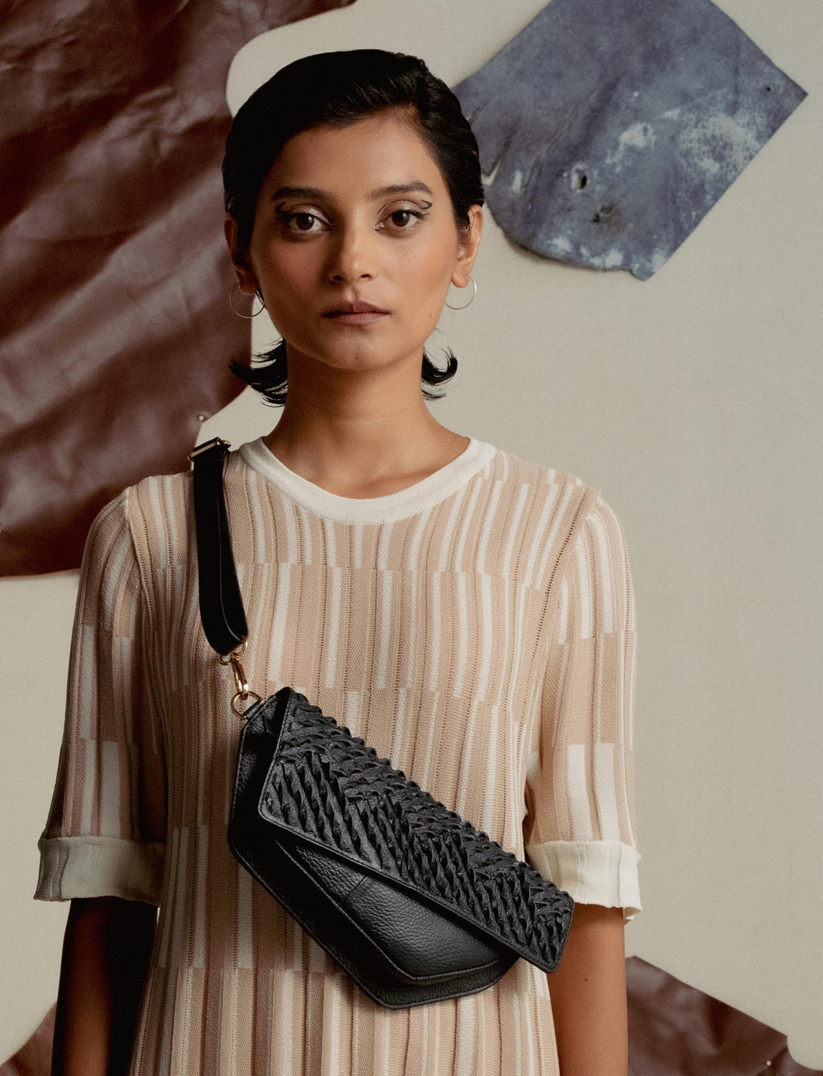 Woman wearing beige striped dress with black woven leather crossbody bag against abstract wall art