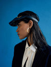 Side profile of woman wearing black leather visor hat with gold embroidery and gold hoop earring against blue background