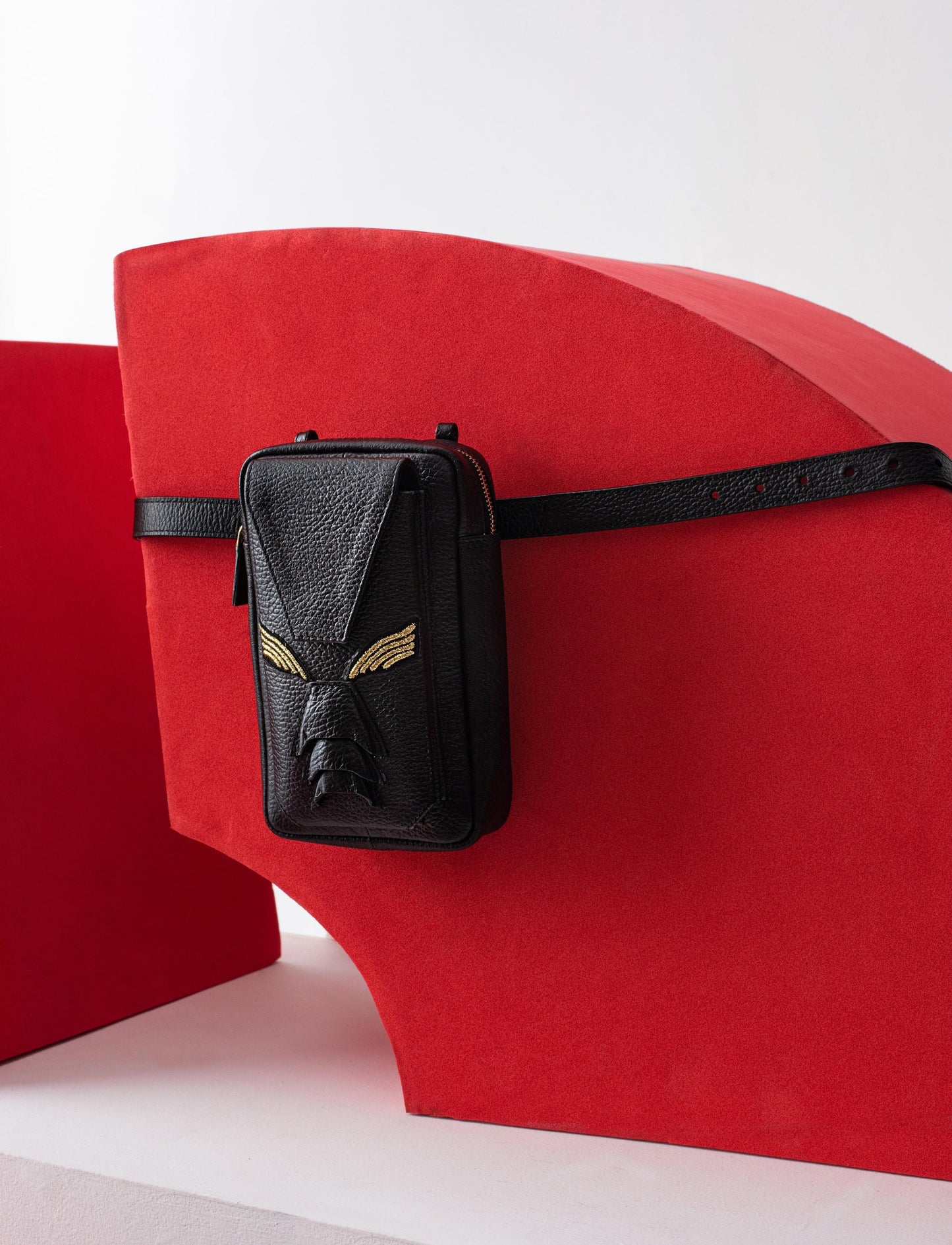 Black leather belt bag with gold wing design on red abstract curved backdrop
