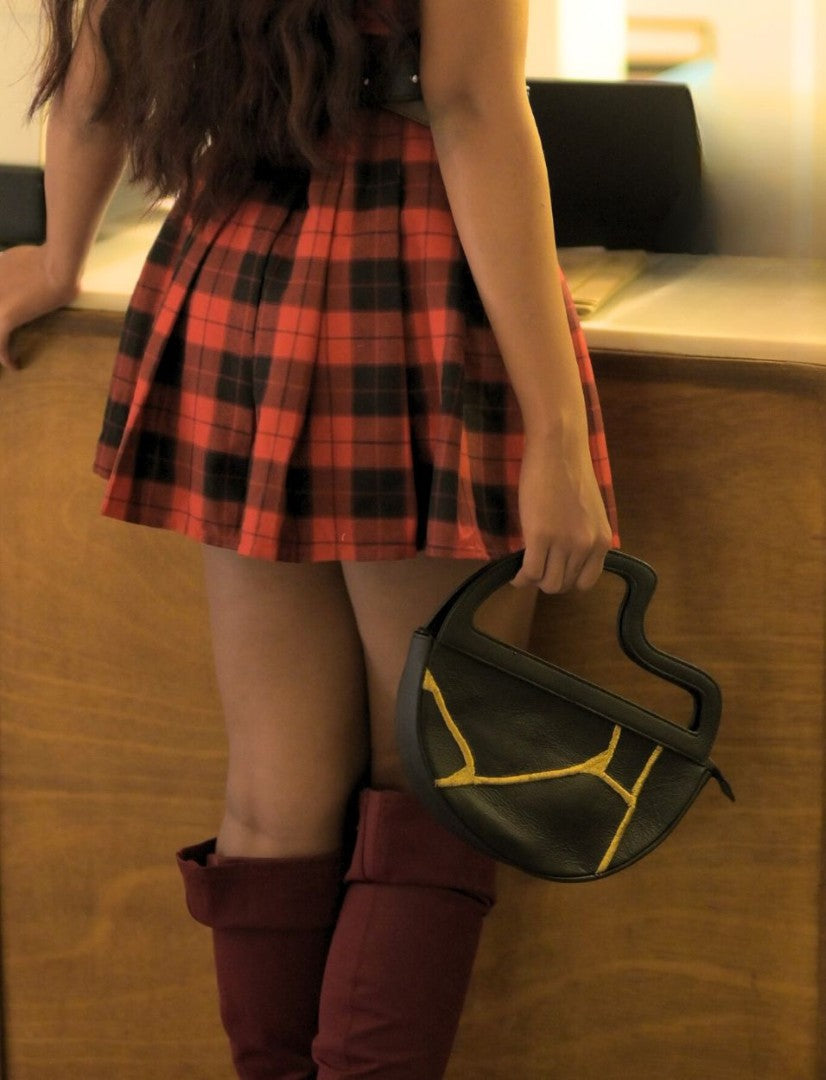 Woman wearing red plaid skirt and maroon boots holding black leather clutch bag with yellow stitching