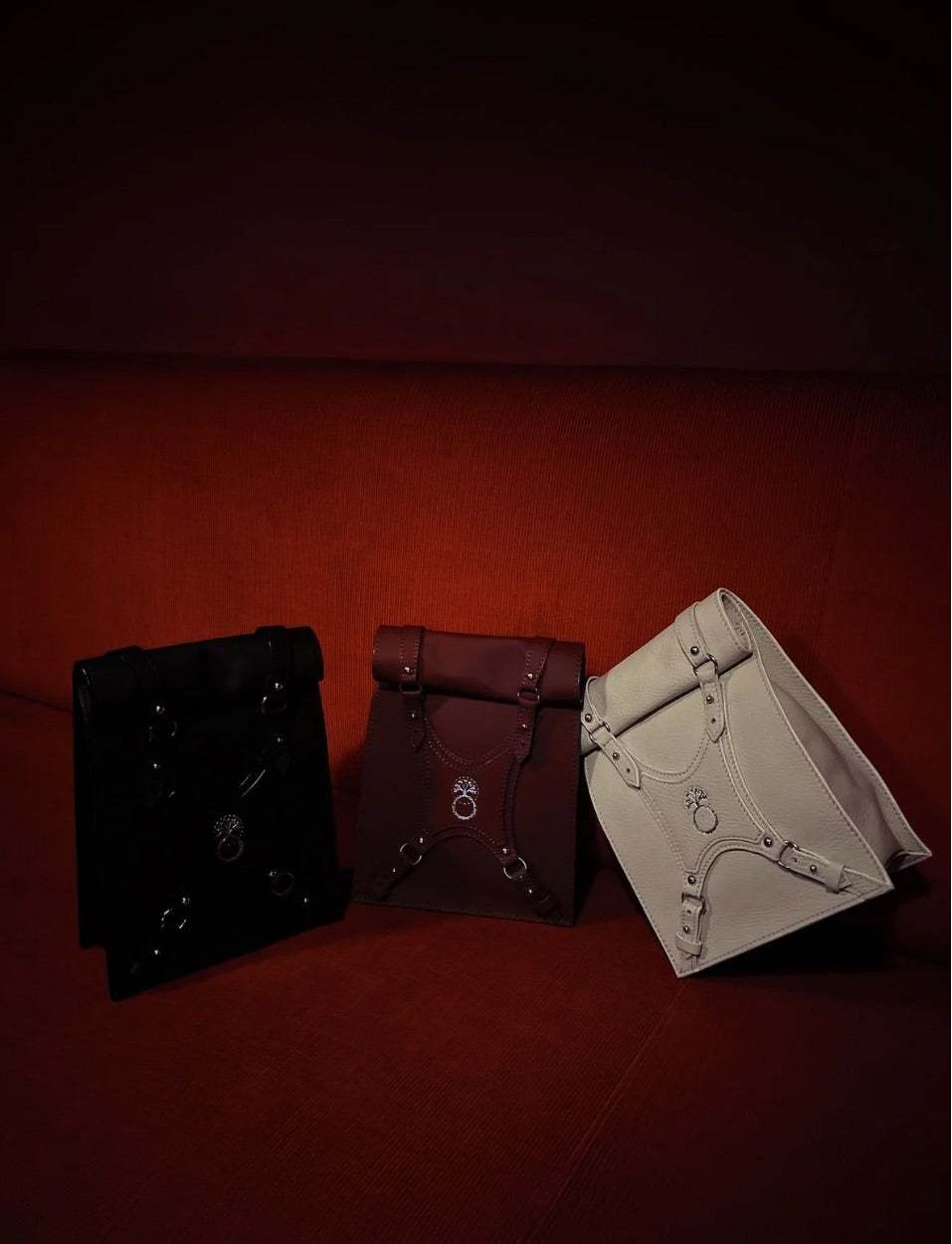 Three leather roll-top backpacks in black, brown, and white resting on a red fabric sofa