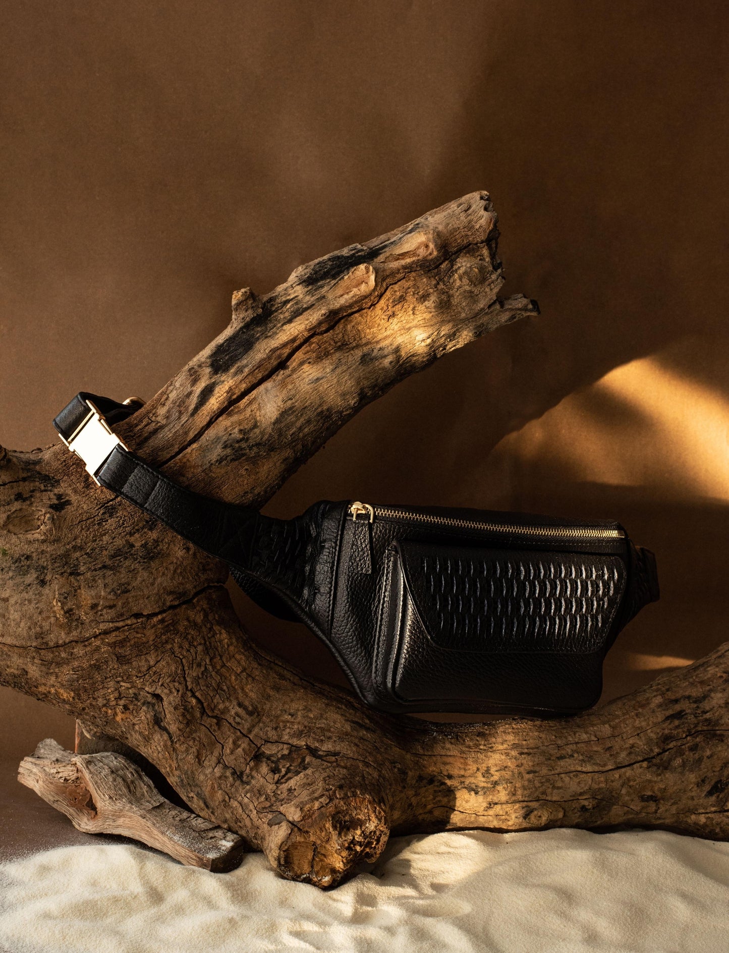 Black textured leather waist bag with gold zipper displayed on rustic driftwood against brown background