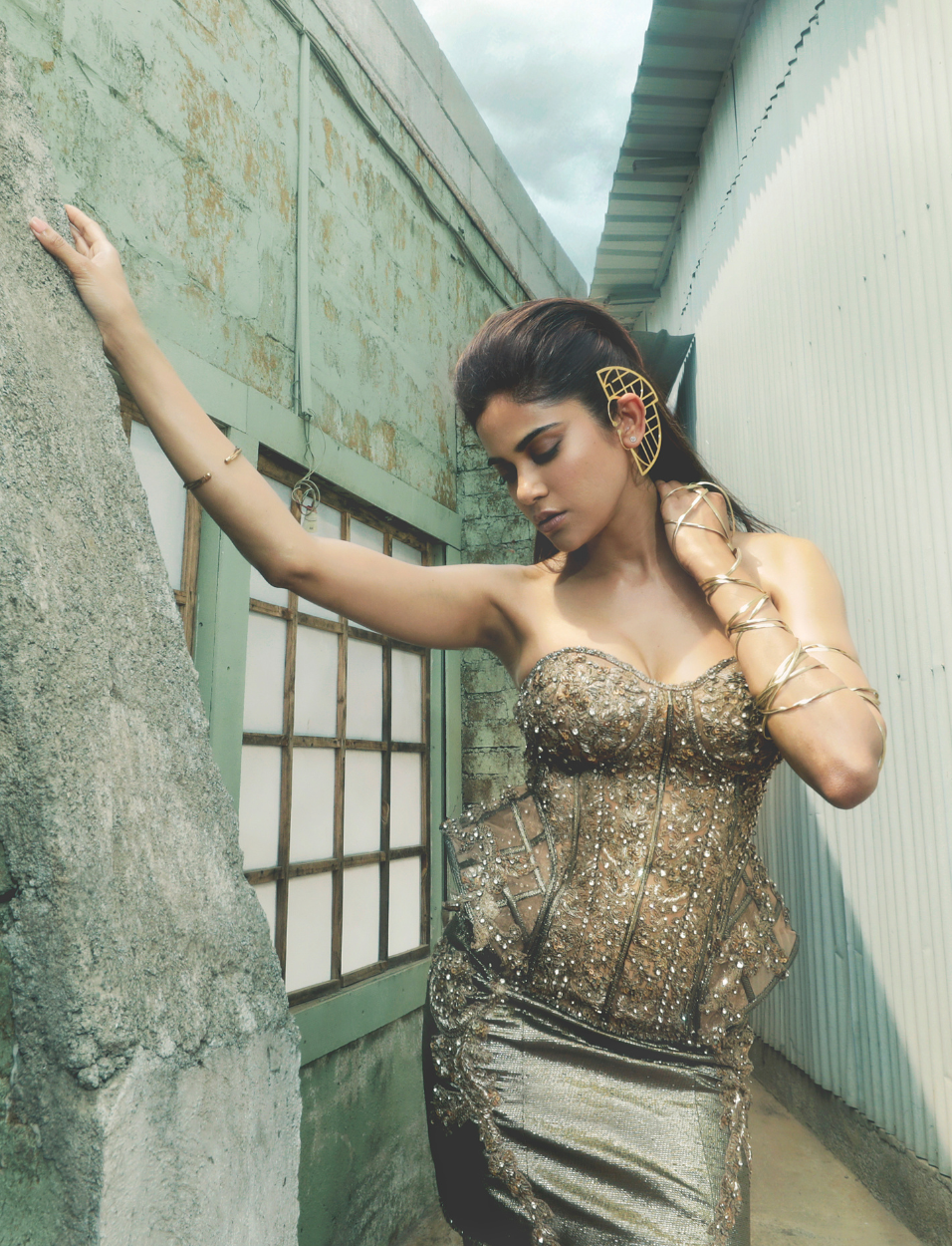 Woman in a sparkly gold corset dress and gold accessories posing against rustic wall and window