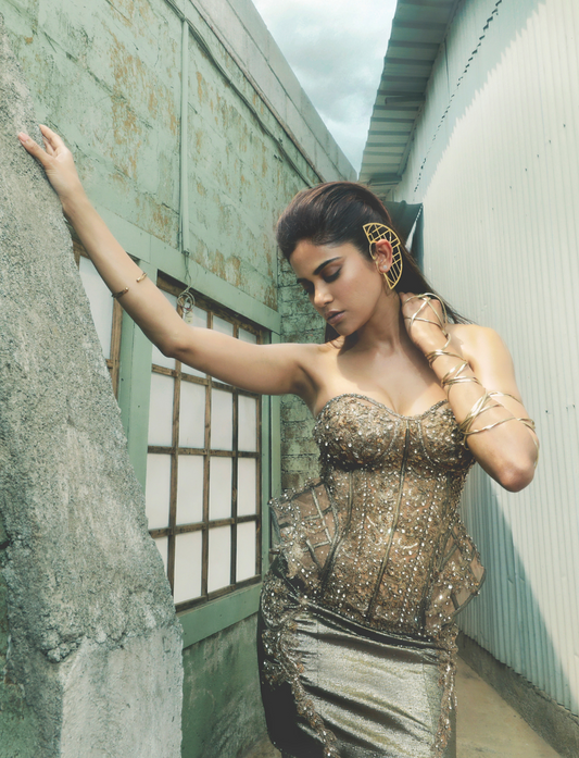 Woman in a sparkly gold corset dress and gold accessories posing against rustic wall and window