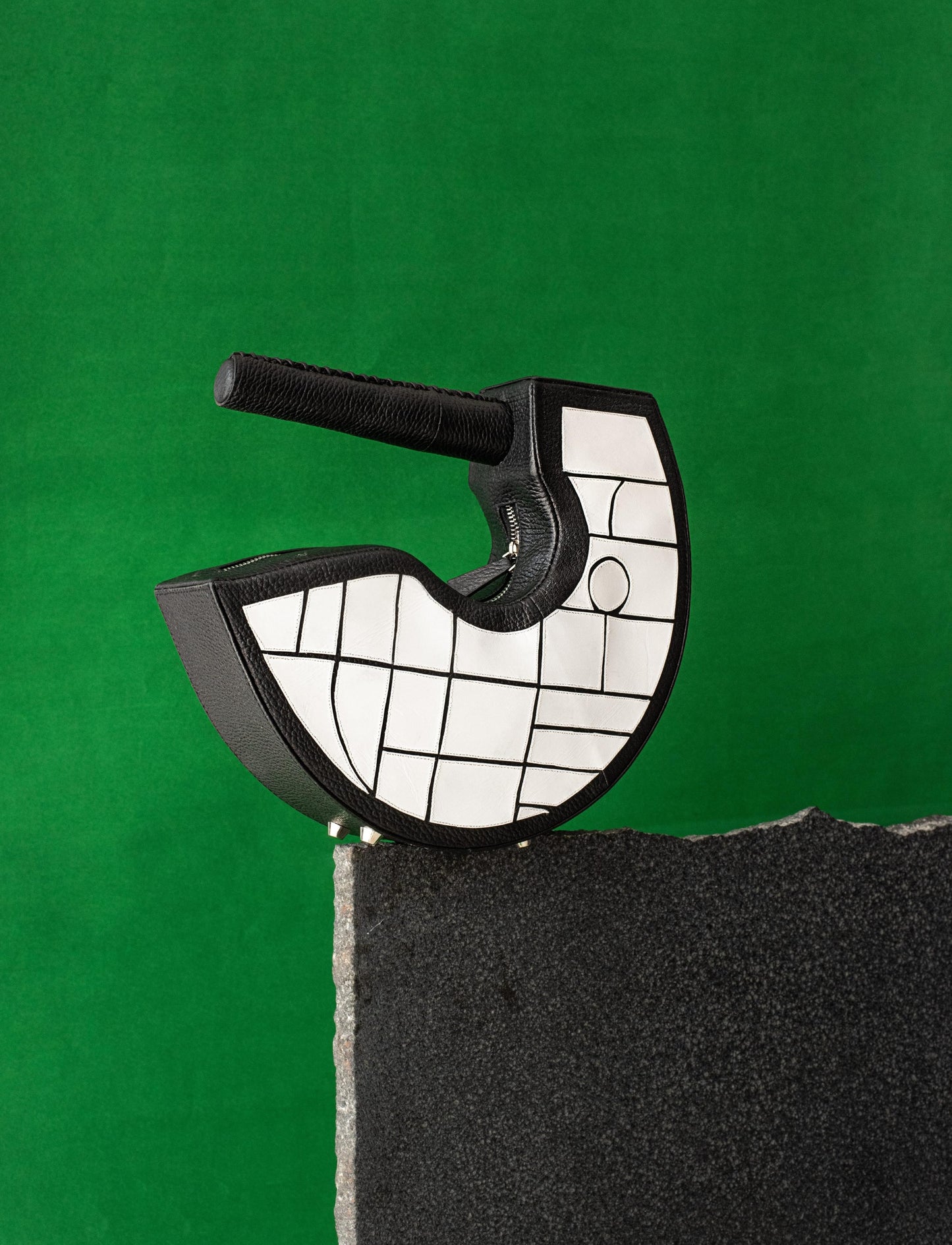 Black and white mosaic leather handbag with circular handle on stone ledge against green background