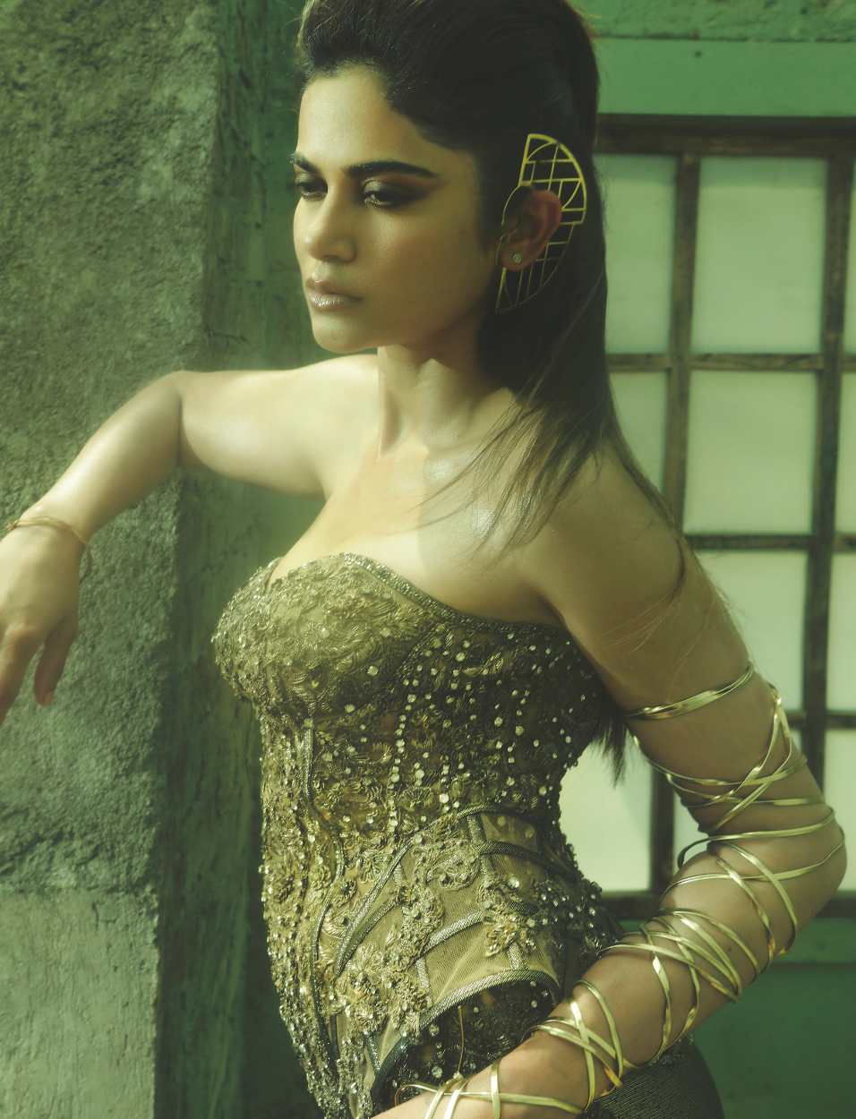 Woman wearing a detailed gold embroidered corset with gold arm wraps and geometric ear accessory in soft light