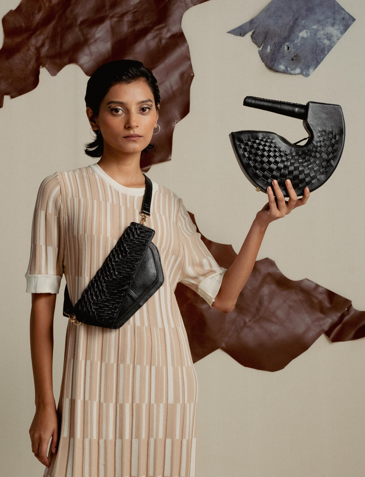 Woman in beige striped dress holding and wearing black woven leather bags against neutral backdrop with leather pieces