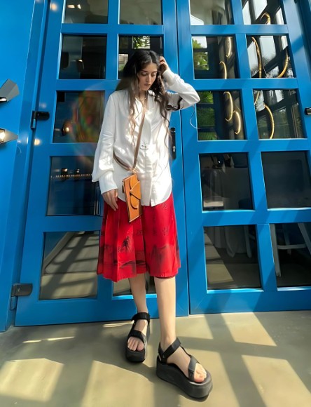 woman in white oversized shirt and red skirt wearing tan leather crossbody bag standing in front of blue glass door