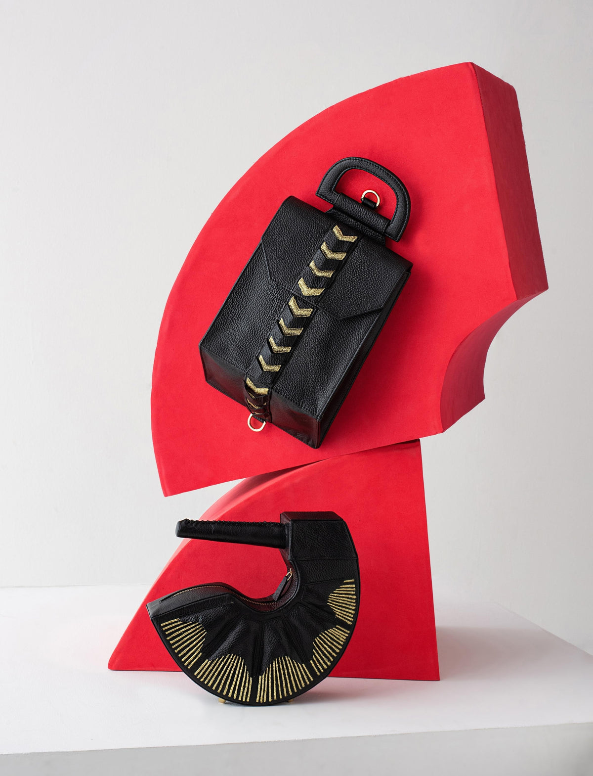 Two black textured handbags with gold accents displayed on a bold red sculptural stand