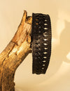 Black woven leather bracelet displayed on a textured tree branch against beige background