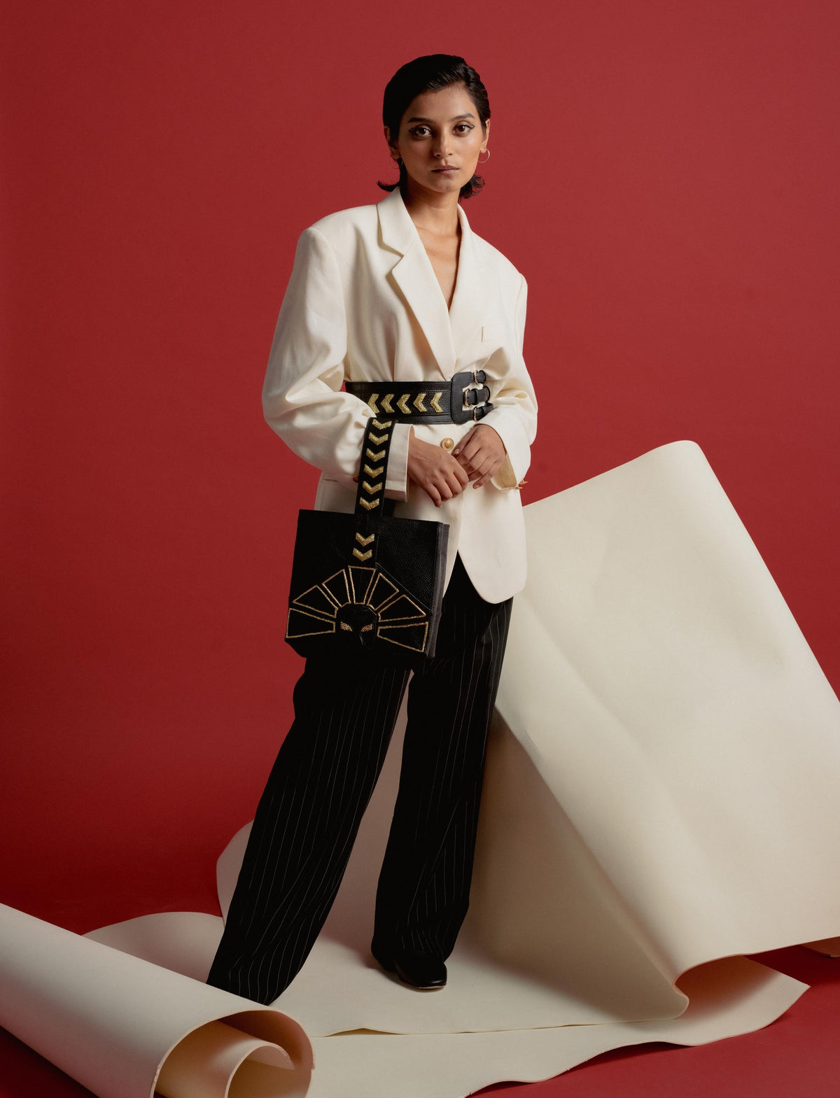 Woman in oversized white blazer and black pinstripe pants holding black bag with gold design on red background