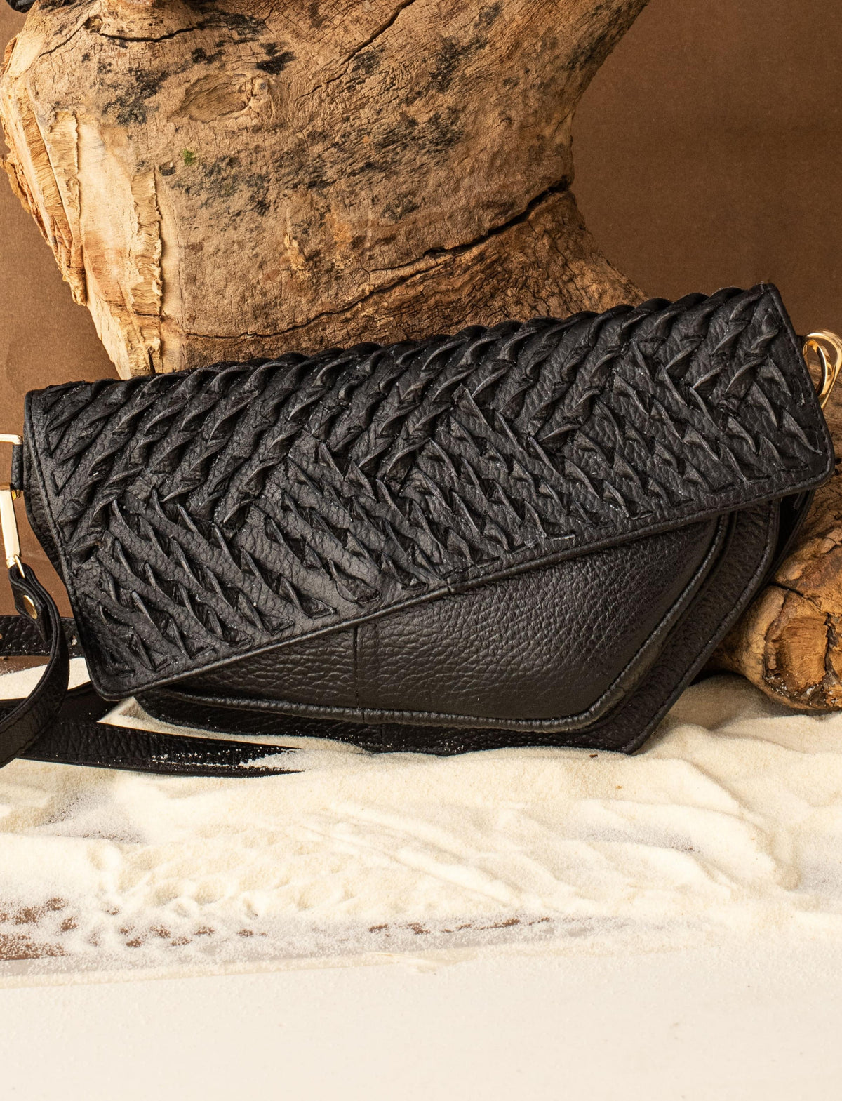 Black textured leather clutch bag with braided flap on sand and wood background