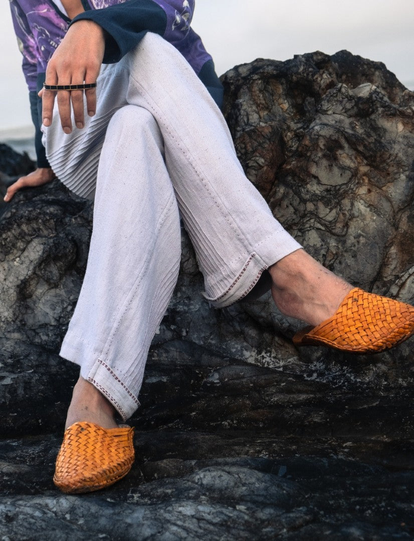 Person sitting on rocky surface wearing white pants and orange woven slip-on shoes, showing hand with ring