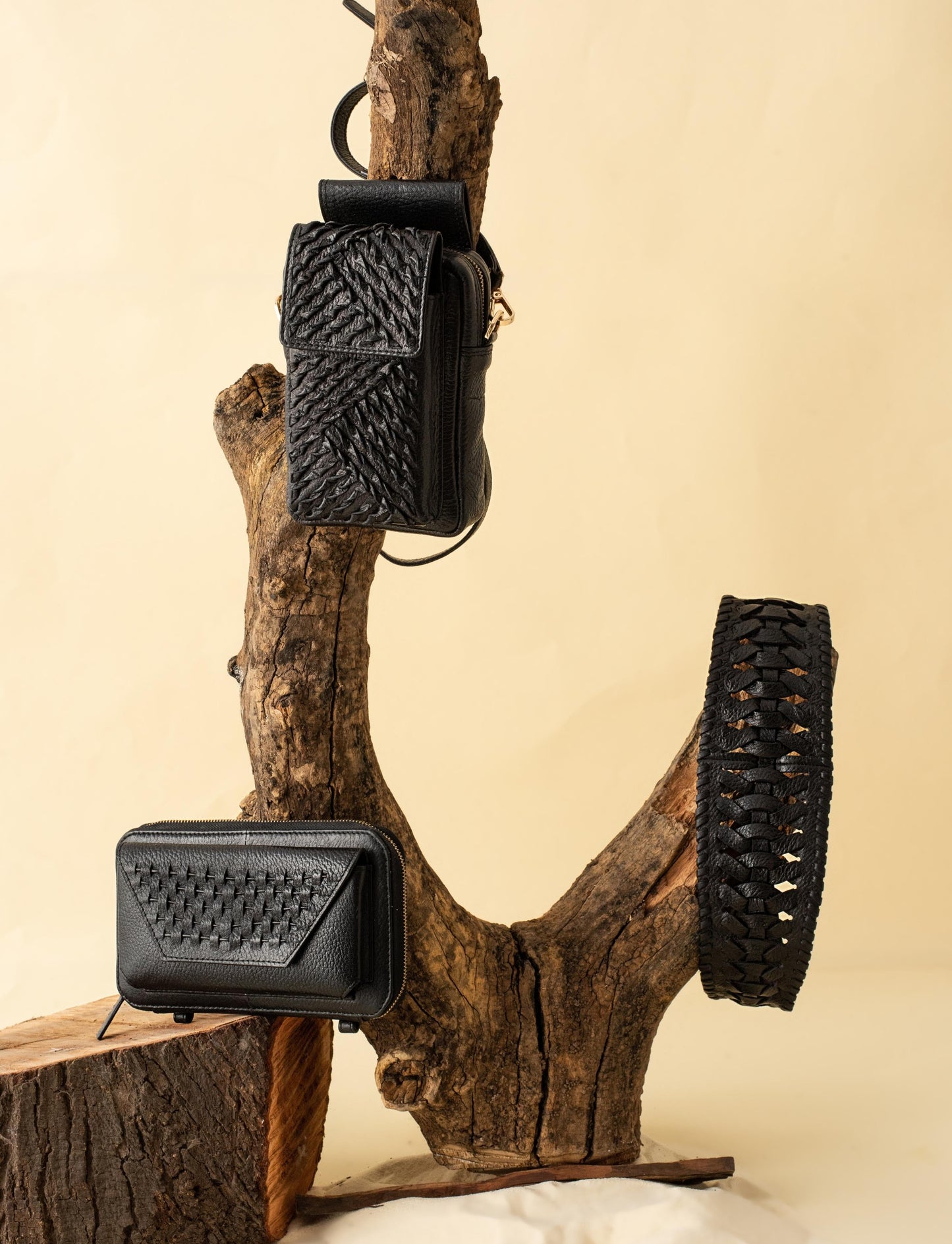 Black woven leather crossbody bag, wallet, and belt displayed on rustic tree branch with beige background