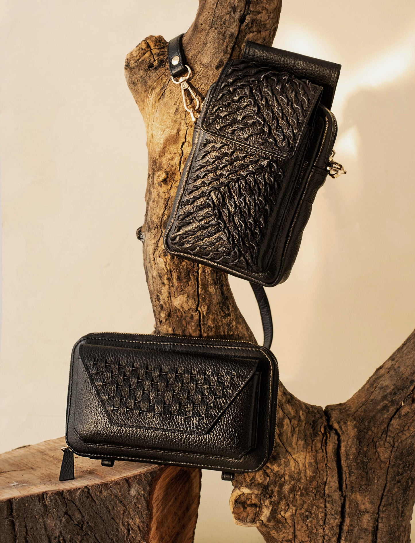 Two black textured leather crossbody bags displayed on natural wooden branches