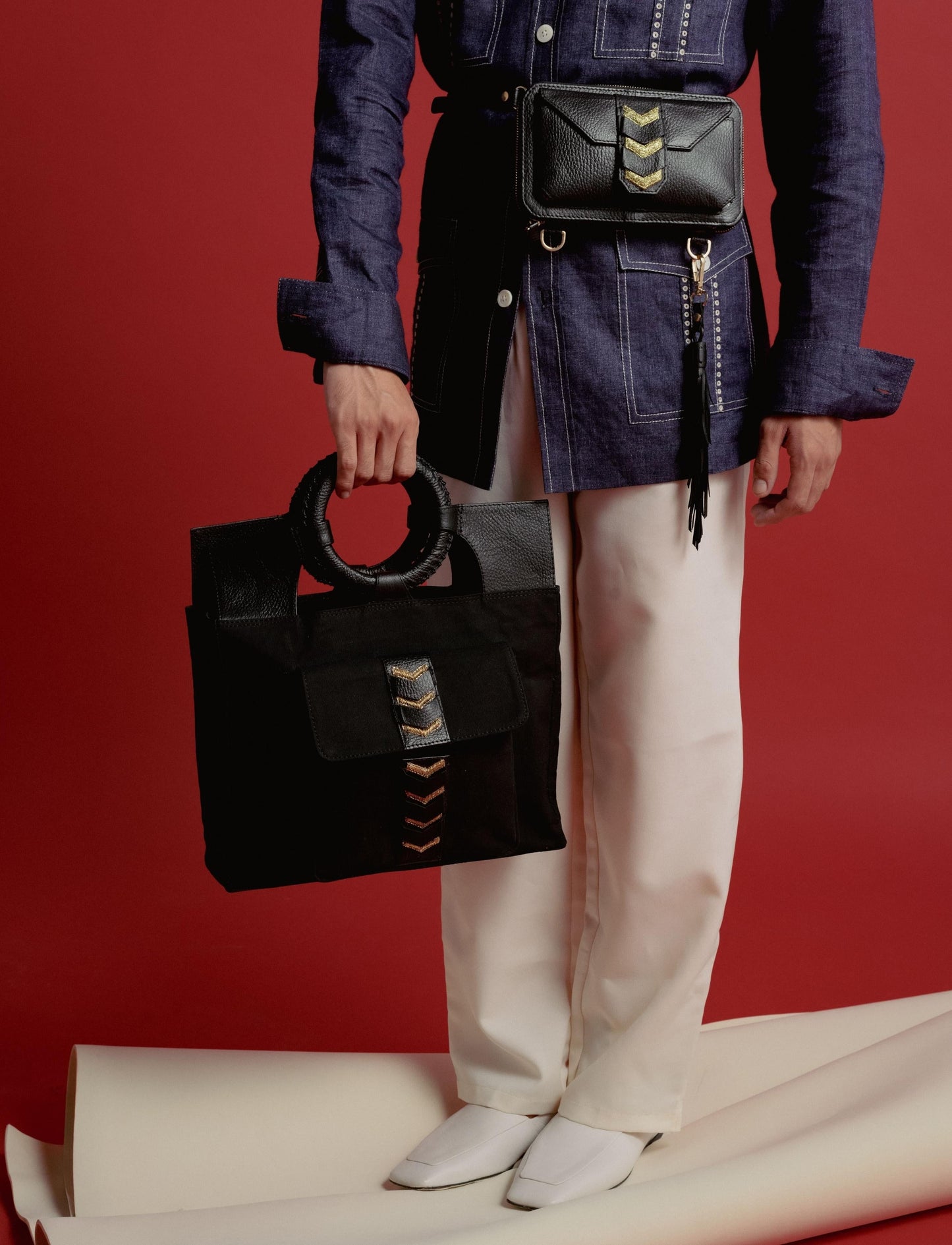 Person in denim jacket and cream pants holding black handbag with circular handle and waist bag on red background