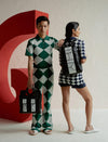 Two models in green and navy harlequin patterned outfits posing with black and white designer bags against a red sculpture.
