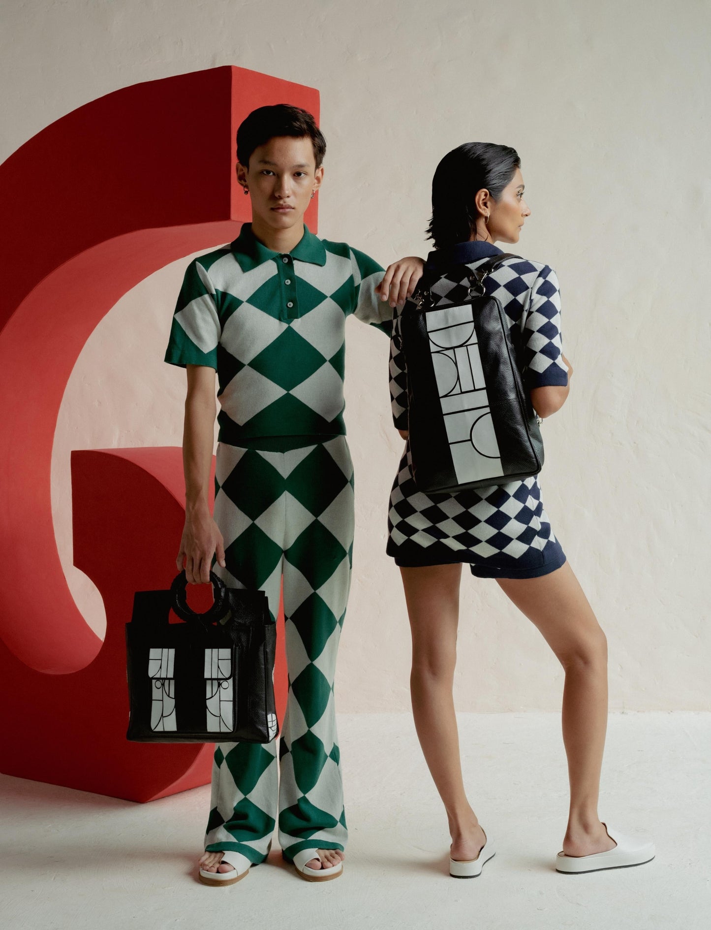 Two models in green and navy harlequin patterned outfits posing with black and white designer bags against a red sculpture.