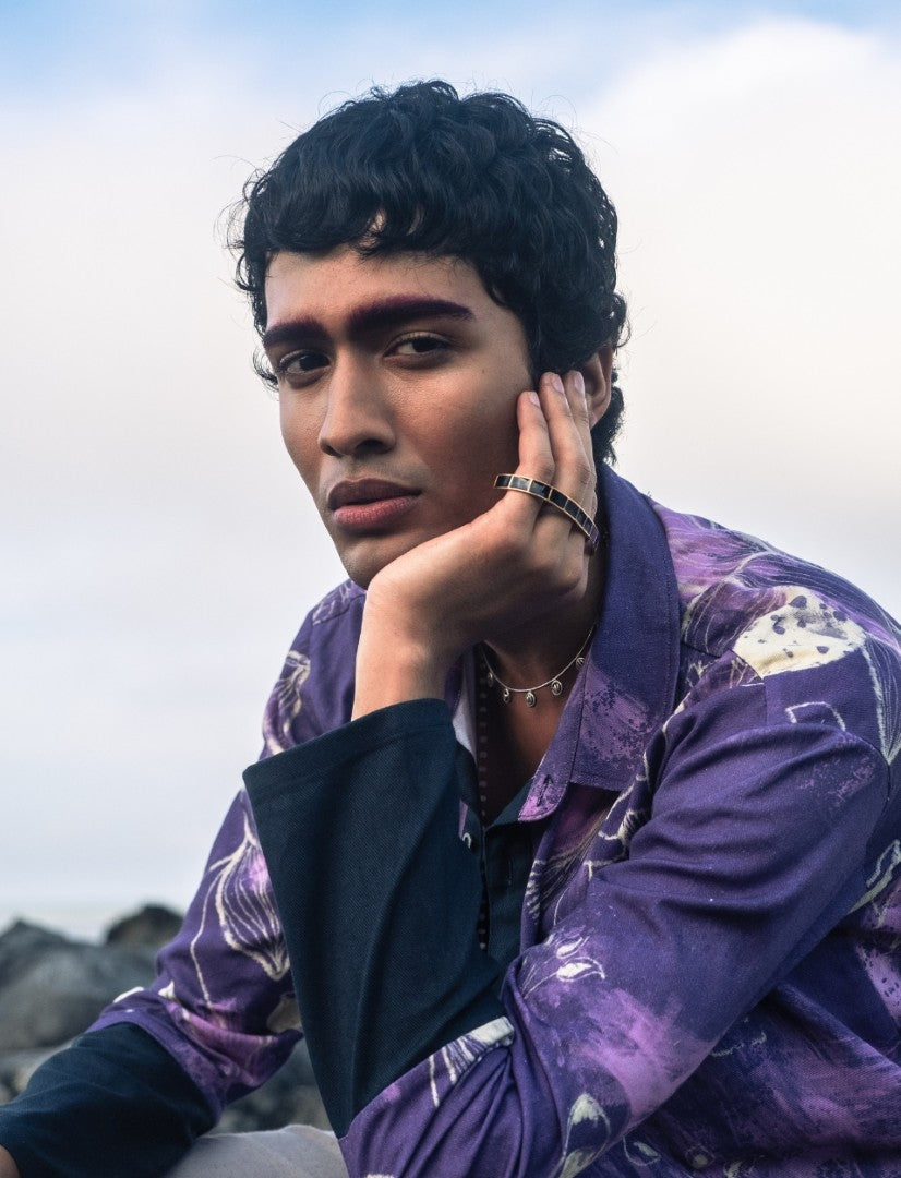 Young man with curly hair wearing a purple patterned jacket and jewelry, resting his face on his hand outdoors