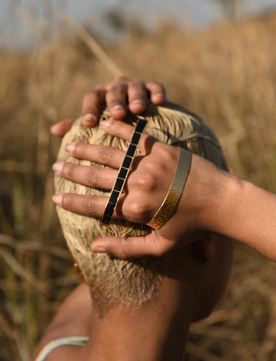 Close-up of a hand with gold and black bracelets on short blonde hair against a blurred outdoor background
