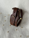 Brown leather bracelet with metal stud and gold accent on white fabric background with scattered small gold beads