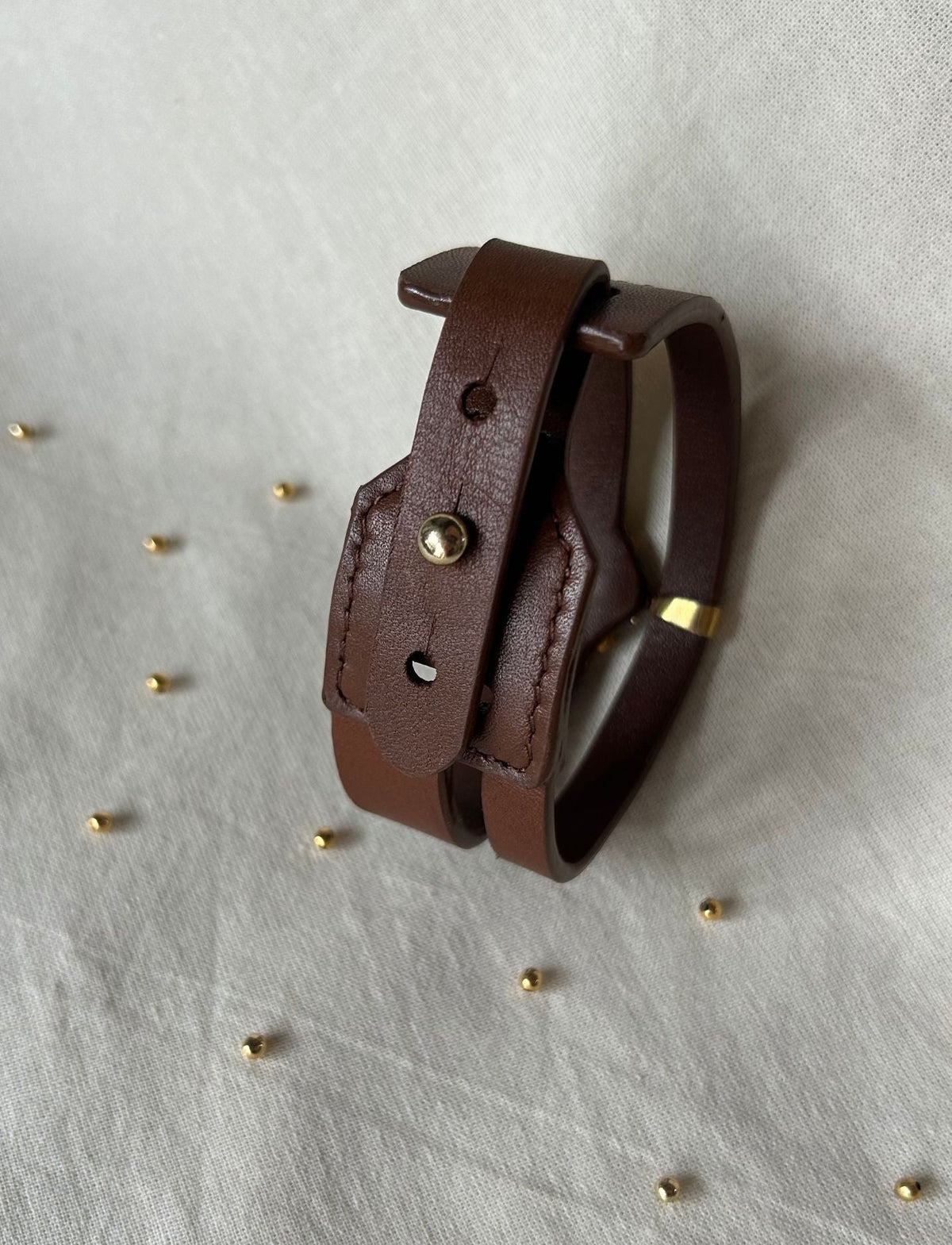Brown leather bracelet with metal stud and gold accent on white fabric background with scattered small gold beads