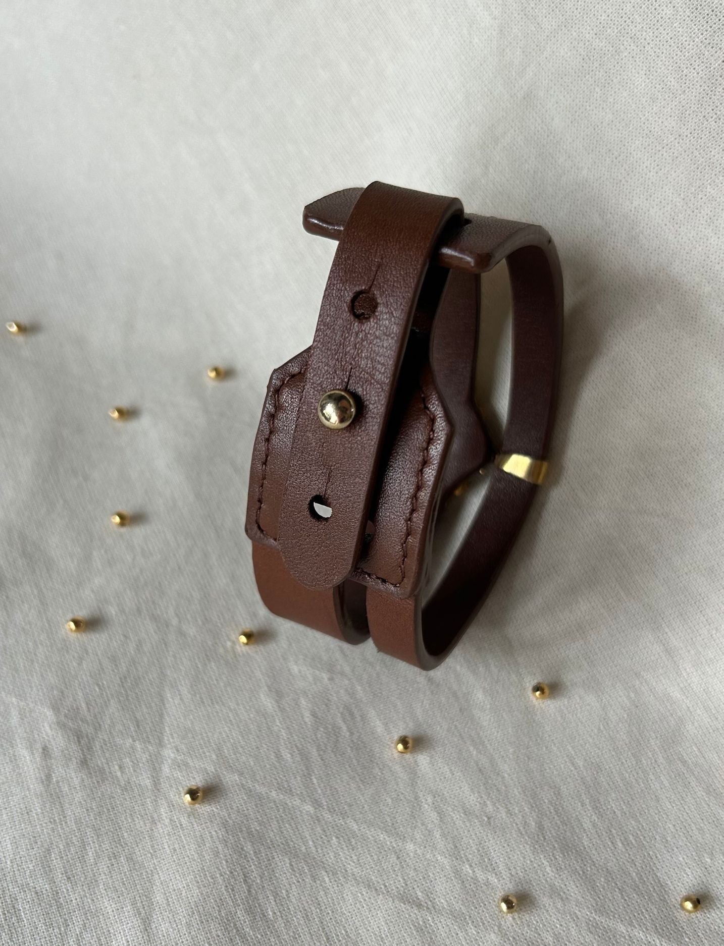 Brown leather bracelet with metal stud and gold accent on white fabric background with scattered small gold beads