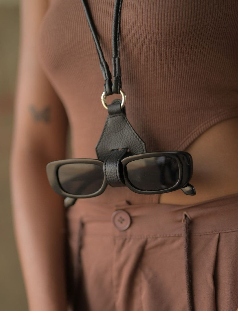Black rectangular sunglasses secured on a black leather protective case hanging from neck on a person wearing brown top and pants