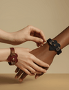 Two hands wearing leather flower bracelets, one in black and one in dark red, on a beige background